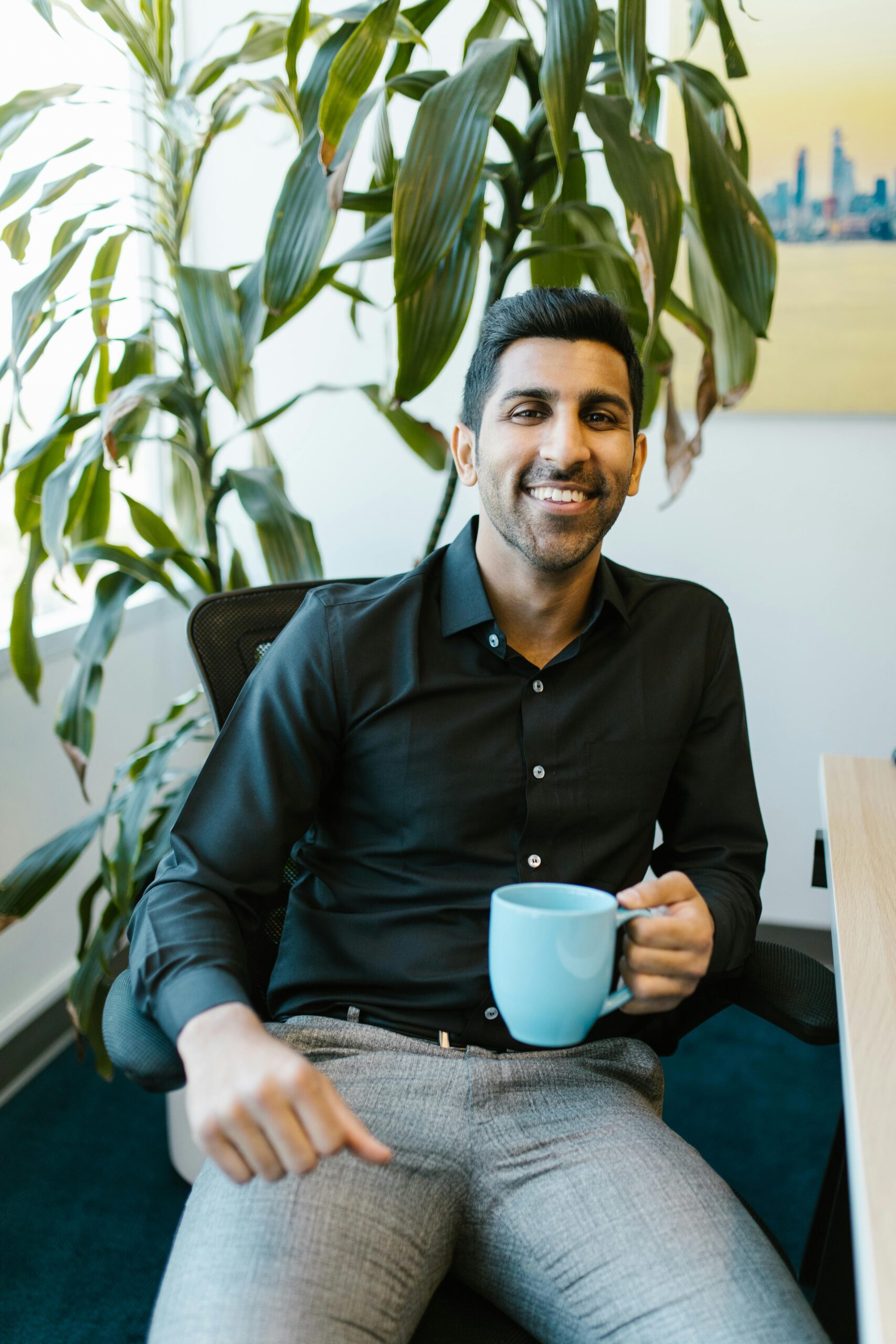 Cheerful man with coffee mug sitting in modern office with plant.